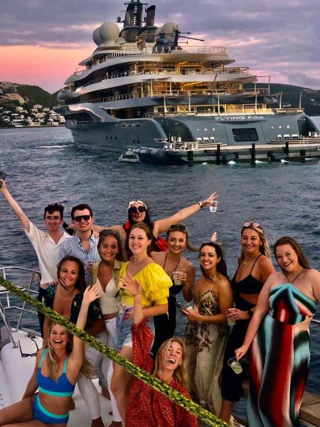 Guests leaning on the bow railing of Sail The Phoenix at sunset with another yacht in view